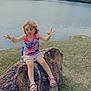 child, girl, rock, lake, water_fountain, grass, trees, outdoor, nature, sandals, colorful_clothing, curly_hair, playful, expression, summer, daylight, sitting, water, scenic, fun