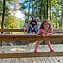 child, children, girl, playful, curly_hair, lollipop, wooden_deck, wooden_railing, bench, outdoor, trees, forest, nature, sunlight, casual_clothing, smiling, happy, friends, daytime, park