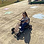 child, go_kart, outdoor, playground, pavement, sunny, person, curly_hair, casual_clothing, jeans, sneakers, adult, umbrella, bench, shadow, daylight, fun, activity, recreation, park