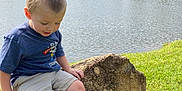 Wyatt is registered to the contest to win money with this photo: child, boy, rock, lake, fountain, water, grass, trees, outdoor, nature, casual_clothing, shorts, tshirt, footwear, hiking_boots, smiling, sitting, sunny, daytime, scenic