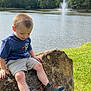 child, boy, rock, lake, fountain, water, grass, trees, outdoor, nature, casual_clothing, shorts, tshirt, footwear, hiking_boots, smiling, sitting, sunny, daytime, scenic