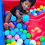 Daniela is registered to the contest to win money with this photo: child, toddler, ball_pit, afro_hair, colorful, play, toy, slide, smile, outdoor, plastic_balls, red_slide, black_clothing, socks, grass, mesh_fence, happy, person, fun, playground