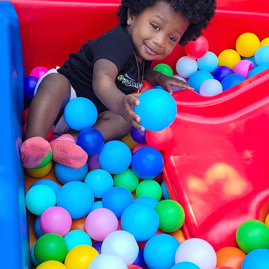 Daniela is registered to the contest to win money with this photo: afro_hair, ball_pit, black_clothing, child, colorful, fun, grass, happy, mesh_fence, outdoor, person, plastic_balls, play, playground, red_slide, slide, smile, socks, toddler, toy
