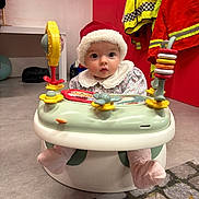 Lilah is registered to the contest to win money with this photo: baby, santa_hat, baby_seat, toy, indoor, child, cute, curious, floor, clothing, sock, playtime, colorful, red_wall, yellow_jacket, plastic_toy, face, person, holiday, festive