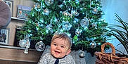 Connor is registered to the contest to win money with this photo: baby, child, christmas_tree, holiday, decorations, ornaments, lights, pajamas, smiling, indoor, wooden_floor, basket, home, festive, happy, seated, person, celebration, seasonal, cozy