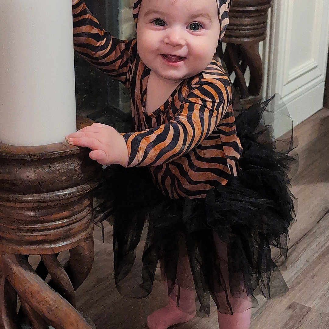 Evelynn is registered to the contest to win money with this photo: baby, black_tutu, bow, candle, child, cute, decor, flooring, happy, headband, holding, home, indoor, person, smiling, standing, tiger_striped_clothing, toddler, tutu, wooden_stand