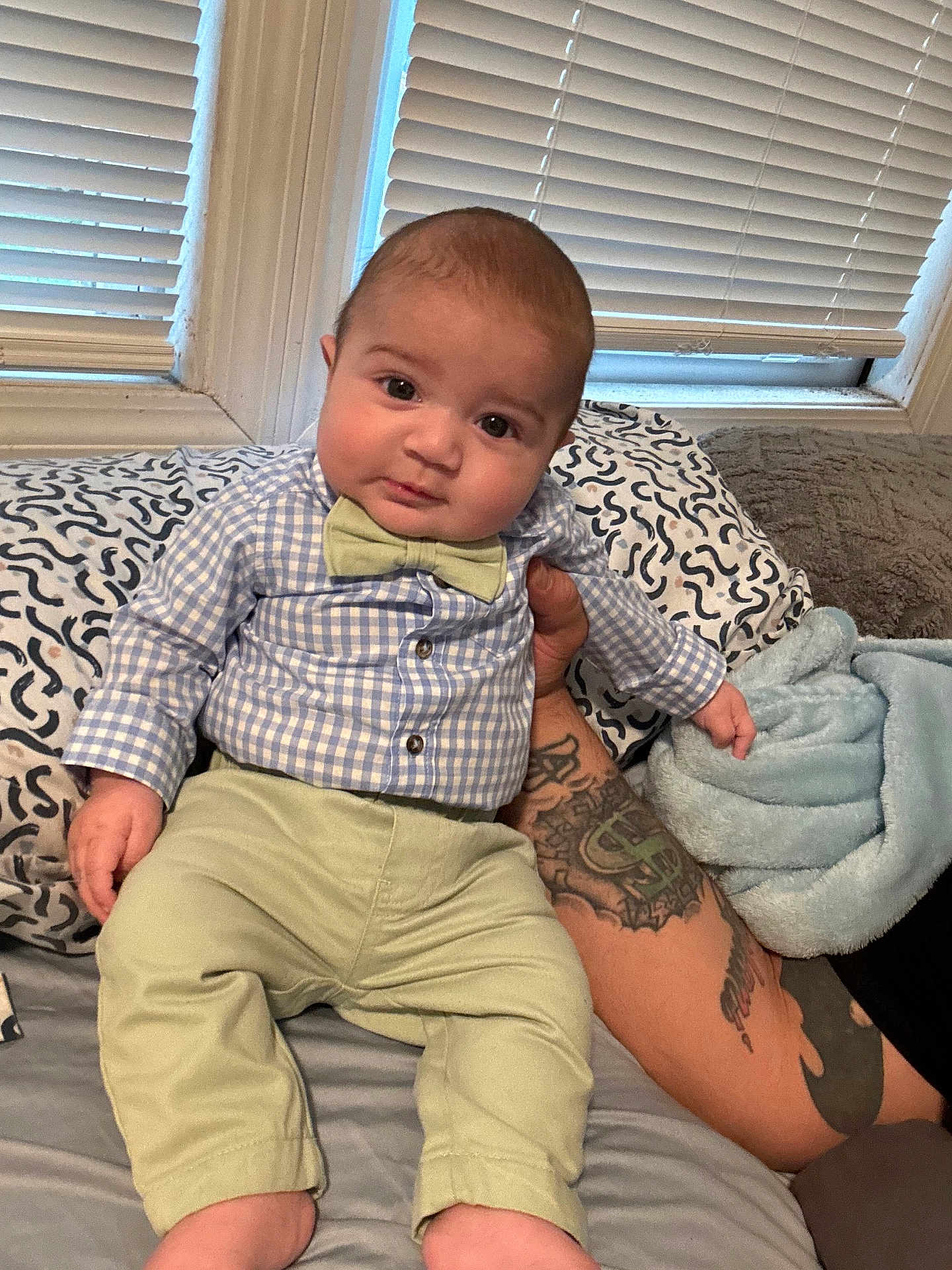 Eric is registered to the contest to win money with this photo: baby, child, infant, bow_tie, tattoo, bed, blanket, window_blinds, shirt, pants, person, indoor, cute, portrait, sitting, hand, face, expression, comfort, home