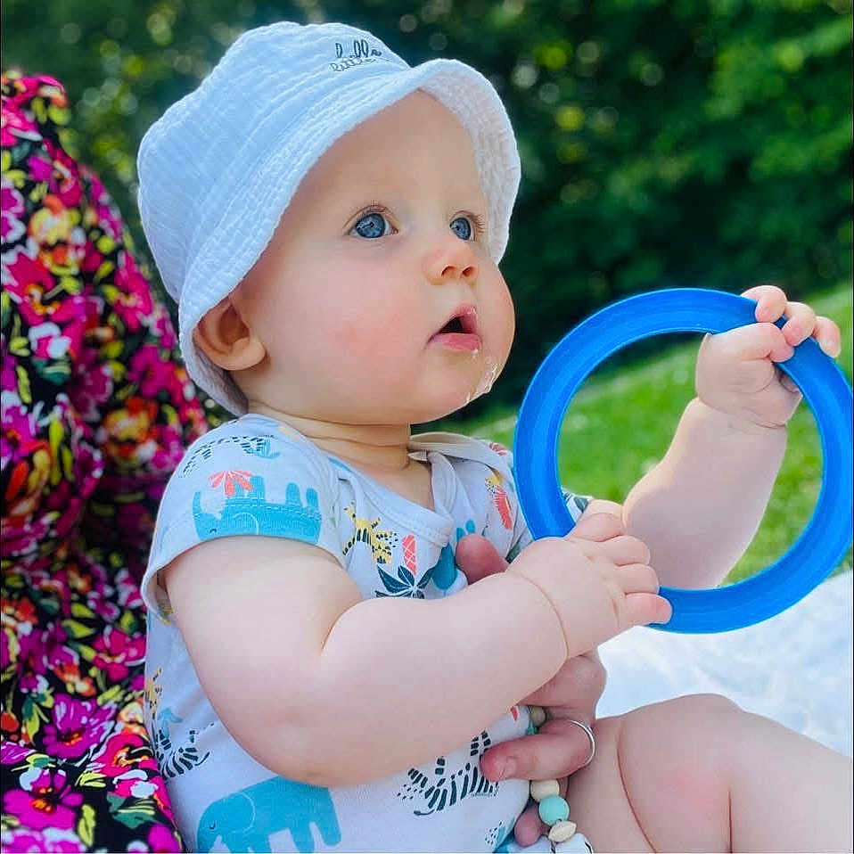 Amaël participe au concours pour gagner de l'argent avec cette photo : baby, blue_eyes, blue_ring, child, colorful_clothing, cute, face, floral_pattern, grass, hand, infant, nature, outdoor, portrait, sitting, skin, summer, sunlight, toy, white_hat
