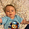 baby, baby_arms, blanket, blue_shirt, casual_clothing, child, colorful, cute, expression, face, headphones, indoor, infant, lying_down, patterned_blanket, person, portrait, smiling, teddy_bear_graphic, young_child