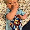 baby, bed, blanket, blue_shirt, casual, child, clothing, curious, cute, face, gesture, hand, indoors, infant, lying_down, patterned_blanket, person, portrait, teddy_bear_graphic, young_child