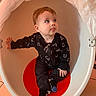 baby, black_clothing, casual_wear, chair, child, cozy, curious, cute, egg_shaped_chair, expression, floor_tiles, indoor, looking_up, playful, portrait, red_seat, sitting, toddler, tongue_out, young_child