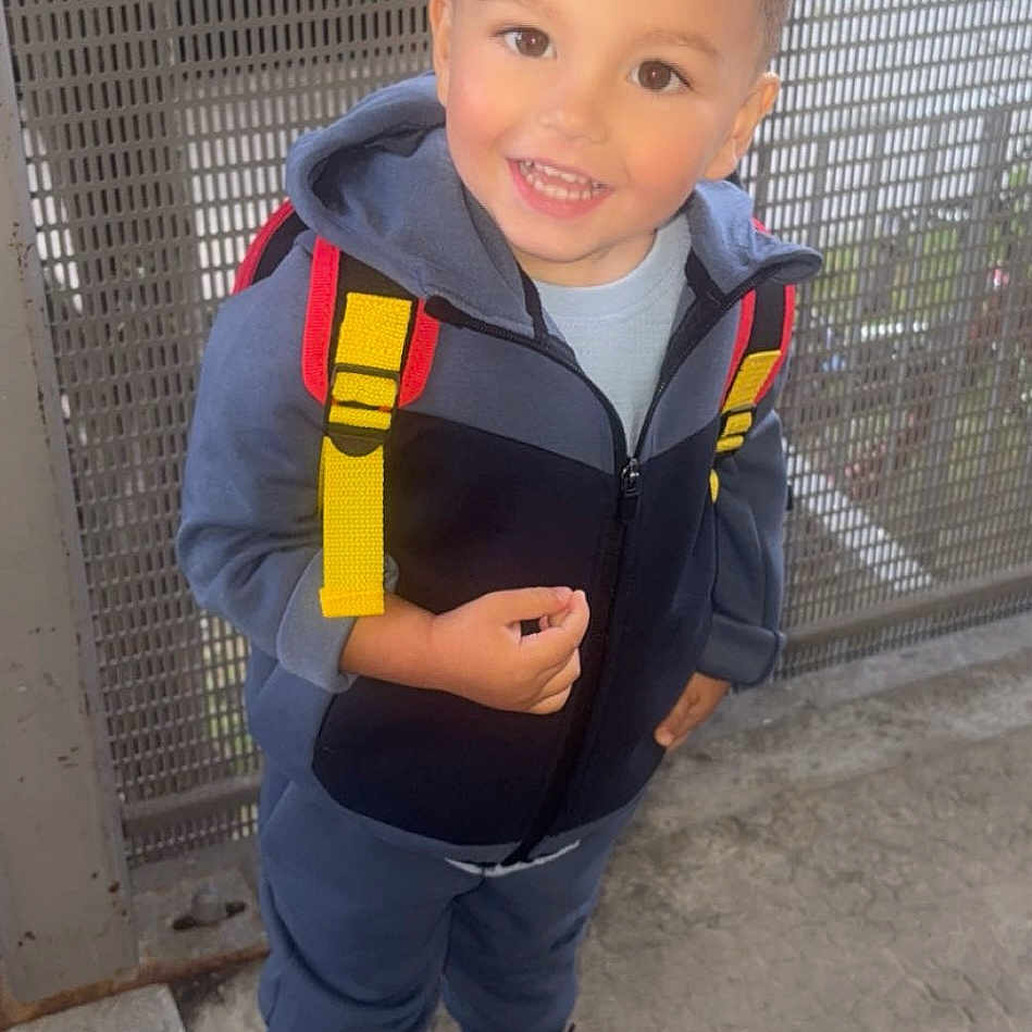 Maloë participe au concours pour gagner de l'argent avec cette photo : backpack, black_shoes, casual_clothing, child, concrete_floor, cute, daylight, full_body, happy, hoodie, metal_fence, outdoor, person, portrait, short_hair, smiling, standing, urban, yellow_straps, young_child