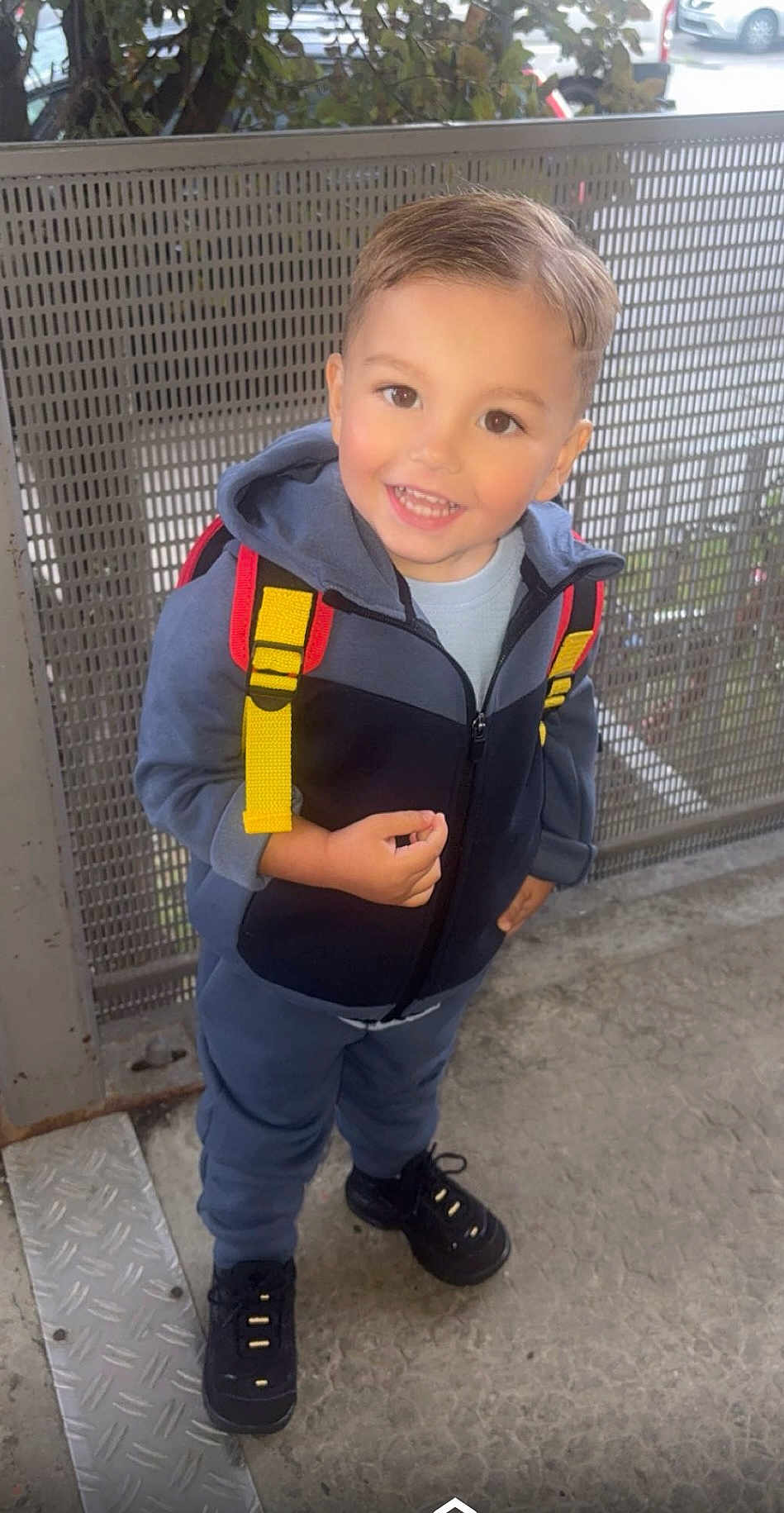 Maloë participe au concours pour gagner de l'argent avec cette photo : child, smiling, hoodie, backpack, yellow_straps, black_shoes, concrete_floor, metal_fence, outdoor, happy, person, casual_clothing, cute, young_child, standing, short_hair, daylight, urban, full_body, portrait