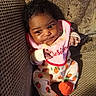 baby, bib, child, clothing, couch, curly_hair, cute, fabric, face, footwear, hand, indoor, infant, orange, patterned_clothing, portrait, seated, smiling, strawberry, textile