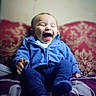 baby, infant, child, laughing, smiling, sitting, blue_clothing, pajamas, bed, bedspread, patterned_background, indoor, portrait, close_up, face, eyes_closed, open_mouth, happy, cozy, photograph