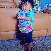 Lucas is registered to the contest to win money with this photo: toddler, child, curly_hair, standing, indoors, living_room, sofa, couch, carpet, colorful_shirt, cartoon, pants, shoes, portrait, full_body, looking_down, hands, furniture, home, casual_clothing