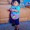 toddler, child, curly_hair, standing, indoors, living_room, sofa, couch, carpet, colorful_shirt, cartoon, pants, shoes, portrait, full_body, looking_down, hands, furniture, home, casual_clothing