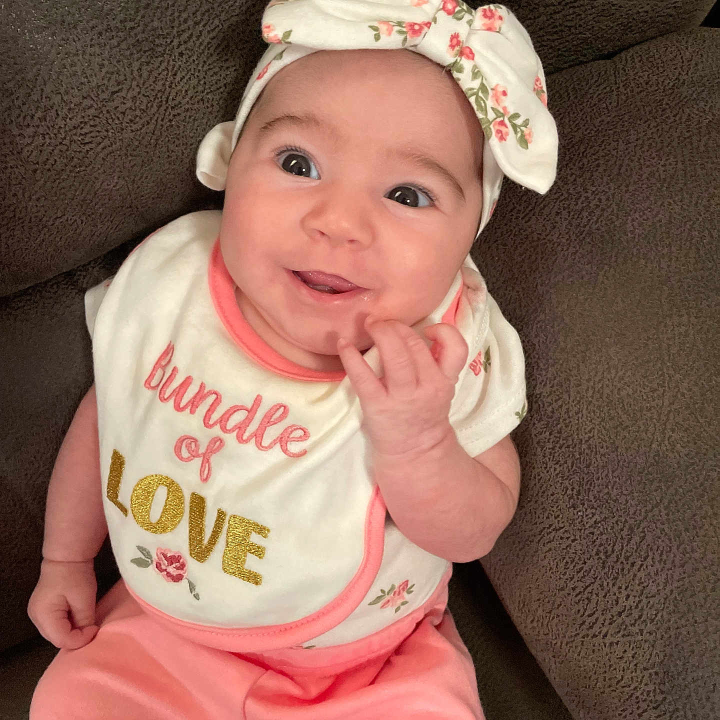Semaj is registered to the contest to win money with this photo: baby, bib, brown_couch, child, clothing, couch, cute, face, floral_pattern, hand, happy, headband, indoors, infant, person, pink_pants, portrait, sitting, smiling, young_child