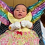 baby, infant, colorful_wings, rainbow, mittens, floral_skirt, yellow_top, indoor, person, newborn, cute, sleepy, patterned_fabric, soft_lighting, cabinet, tabletop, decor, hospital_room, resting, portrait