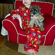 Winston is registered to the contest to win money with this photo: toddler, child, dog, puppy, red_pajamas, armchair, carpet, living_room, cushion, wooden_wall, family_photos, table, smiling, barefoot, indoor, cute, pet, cozy, home, furniture
