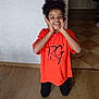 child, smile, kneeling, orange_tshirt, black_pants, wooden_floor, indoor, joyful, hands_on_face, curly_hair, wall, doorway, tile_floor, light_switch, casual_clothing, portrait, person, home, happy, young
