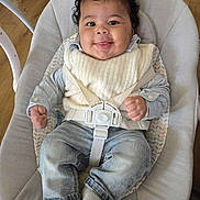 Maylan participe au concours pour gagner de l'argent avec cette photo : baby, baby_seat, child, clothing, curly_hair, cute, furniture, happy, home, indoor, infant, jeans, person, portrait, safety_straps, seated, smiling, socks, vest, wooden_floor