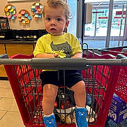 Ronan is registered to the contest to win money with this photo: blue_shoes, child, clothing, curly_hair, door, entrance, expression, face, floor_tiles, indoor, legs, person, retail, shopping, shopping_cart, sticky_notes, store, toddler, window, yellow_shirt