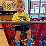 blue_shoes, child, clothing, curly_hair, door, entrance, expression, face, floor_tiles, indoor, legs, person, retail, shopping, shopping_cart, sticky_notes, store, toddler, window, yellow_shirt