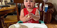Ronan is registered to the contest to win money with this photo: toddler, child, baby, highchair, booster_seat, red_shirt, food, wooden_chair, wooden_table, indoor, living_room, painting, armchair, floor, person, face, hands, feet, hairstyle, expression