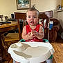 toddler, child, baby, highchair, booster_seat, red_shirt, food, wooden_chair, wooden_table, indoor, living_room, painting, armchair, floor, person, face, hands, feet, hairstyle, expression