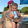 baby, man, tattoos, pool, outdoor, summer, drinkware, cap, sunglasses, child, holding, water, bench, table, trees, sunlight, playful, barefoot, black_swimwear, relaxation