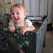 Carsen is registered to the contest to win money with this photo: baby, child, playpen, smile, happy, indoor, curtain, door, carpet, clothing, person, furniture, hand, foot, mesh, pattern, green, yellow, blue, toy