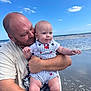 baby, barefoot, beach, casual_clothing, child, clouds, family, happy, holding, man, nature, ocean, outdoor, person, red_crab_pattern, short_sleeve, sky, smile, striped_overalls, water