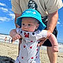 adult_hands, baby, beach, blue_hat, casual_clothing, child, crab_pattern, cute, infant, ocean, outdoor, person, playful, portrait, sand, sky, striped_overalls, summer, sunny, vacation