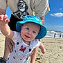 Matthew is registered to the contest to win money with this photo: adult_hand, baby, beach, blue_sky, cartoon_print, child, crocs, ocean, outdoor, overalls, person, reaching_hand, red_crab_pattern, sand, smiling, striped_clothing, sunhat, sunny, tshirt, waves