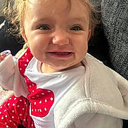 Mayva is registered to the contest to win money with this photo: baby, child, smiling, curly_hair, red_clothing, white_jacket, polka_dot, sitting, indoor, couch, happy, face, person, portrait, closeup, cute, infant, apparel, cozy, young
