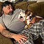 man, dog, boxer_dog, couch, tattoo, plaid_pants, indoor, curtains, relaxing, pet, companionship, casual_clothing, brown_furniture, facial_hair, resting, comfort, animal, human, household, friendship