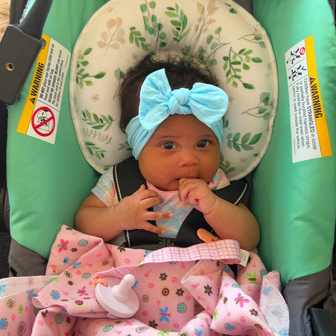 Kohani is registered to the contest to win money with this photo: baby, blanket, blue_bow, car_seat, child, comfort, cute, expression, face, floral_pattern, hand, headband, indoor, infant, looking, person, pink, safety_harness, straps, young