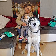 Tiago a rejoint le concours — aidez-le/la à gagner de superbes lots ! woman, child, dog, siberian_husky, couch, indoor, living_room, cityscape_art, casual_clothing, child_shoes, seated, pet, family, home, blanket, pillow, floor, bag, phone, coat