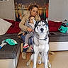 woman, child, dog, siberian_husky, couch, indoor, living_room, cityscape_art, casual_clothing, child_shoes, seated, pet, family, home, blanket, pillow, floor, bag, phone, coat