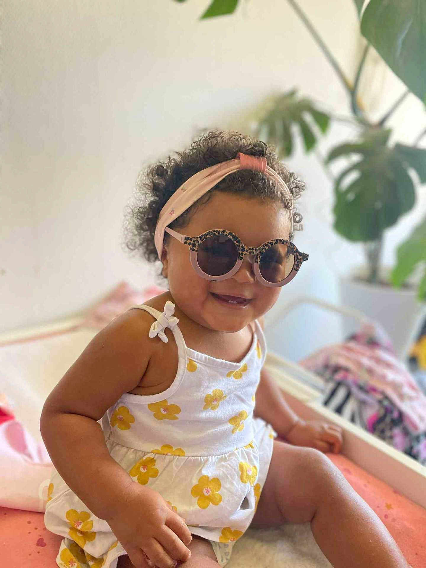 Ariane participe au concours pour gagner de l'argent avec cette photo : toddler, child, sunglasses, headband, curly_hair, dress, flower_pattern, bed, indoor, smile, happy, skin, person, furniture, plant, leafy_plant, pink, yellow, casual, cute