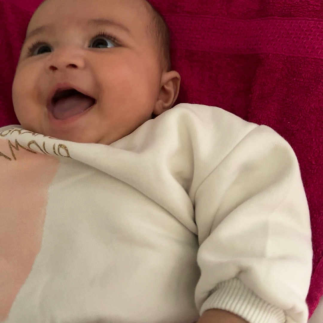 Zeïna a rejoint le concours — aidez-le/la à gagner de superbes lots ! baby, cheerful, child, clothing, cozy, cute, diapers, face, hand, happy, indoors, infant, lying_down, patterned_pants, portrait, red_towel, skin, smiling, soft_fabric, white_sweatshirt