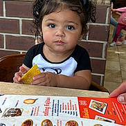 Paula Monica is registered to the contest to win money with this photo: child, toddler, girl, portrait, restaurant, dining, menu, food, tortilla_chip, snack, chair, table, brick_wall, indoor, sitting, curly_hair, hair_bows, looking_at_camera, facial_expression, family
