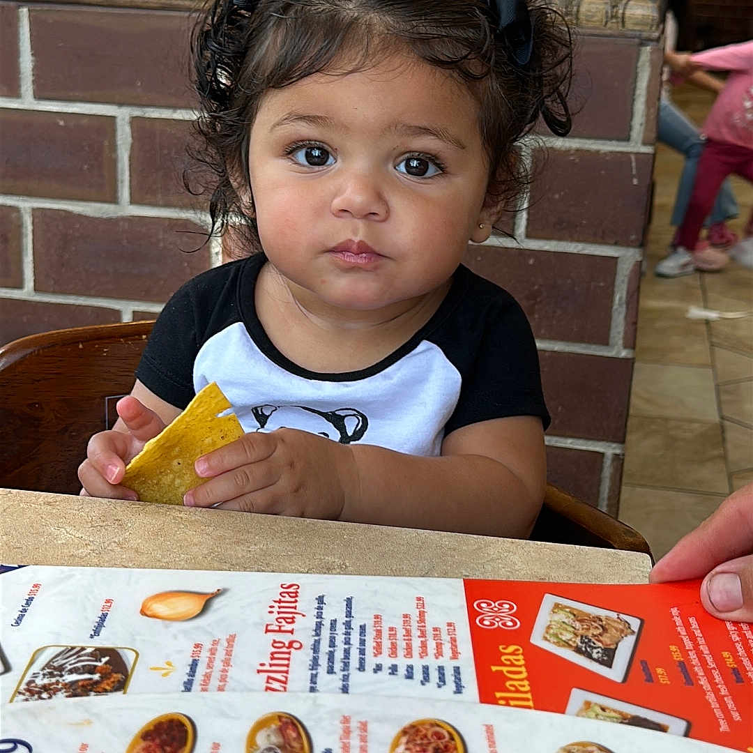 Paula Monica is registered to the contest to win money with this photo: child, toddler, girl, portrait, restaurant, dining, menu, food, tortilla_chip, snack, chair, table, brick_wall, indoor, sitting, curly_hair, hair_bows, looking_at_camera, facial_expression, family