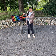 Daniela participe au concours pour gagner de l'argent avec cette photo : child, girl, outdoor, xylophone, musical_instrument, mallets, gravel, trees, stone_wall, leopard_print_jacket, black_pants, sneakers, play, park, nature, greenery, fence, standing, smiling, recreation