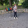 child, girl, outdoor, xylophone, musical_instrument, mallets, gravel, trees, stone_wall, leopard_print_jacket, black_pants, sneakers, play, park, nature, greenery, fence, standing, smiling, recreation