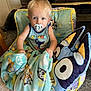 toddler, child, blanket, pacifier, chair, pillow, cartoon_character, indoor, carpet, fireplace, stone_wall, blue_clothing, cute, home, comfort, young_child, sitting, curious, toy, cozy
