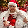 baby, infant, santa_hat, christmas_tree, christmas_lights, presents, holiday, festive, red_clothing, white_clothing, pillow, animal_print, smiling, indoors, celebration, cute, child, seasonal, decorations, happy