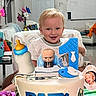 toddler, child, baby, smiling, birthday, birthday_cake, high_chair, toy, doll, baby_bottle, money, party, indoors, living_room, furniture, portrait, blond_hair, blue_shirt, celebration, wrapping_paper
