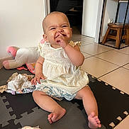 Eliana a rejoint le concours — aidez-le/la à gagner de superbes lots ! baby, child, sitting, play_mat, indoor, white_dress, shorts, toys, floor, happy, smiling, finger_in_nose, cute, infant, footwear_free, home, furniture, wooden_stool, door, tile_floor